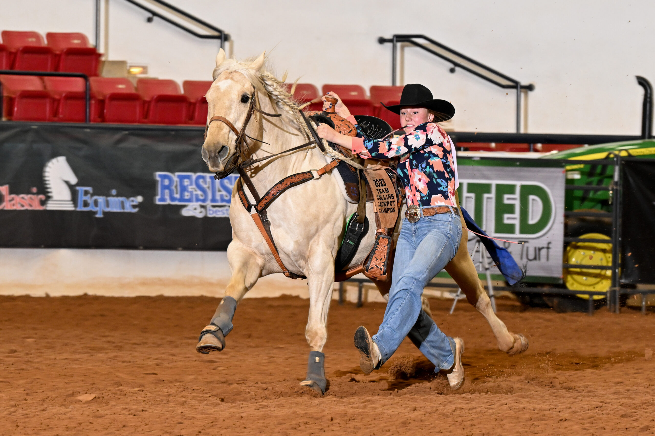 Barrel racer on palomino horse rounds a barrel at indoor rodeo arena with red seating in background