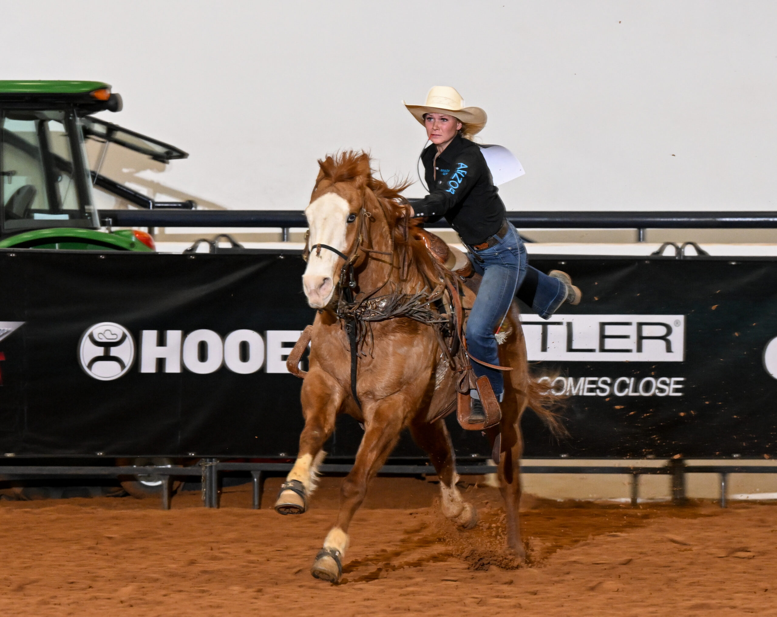 Barrel racer on brown and white horse gallops past sponsor boards at rodeo arena