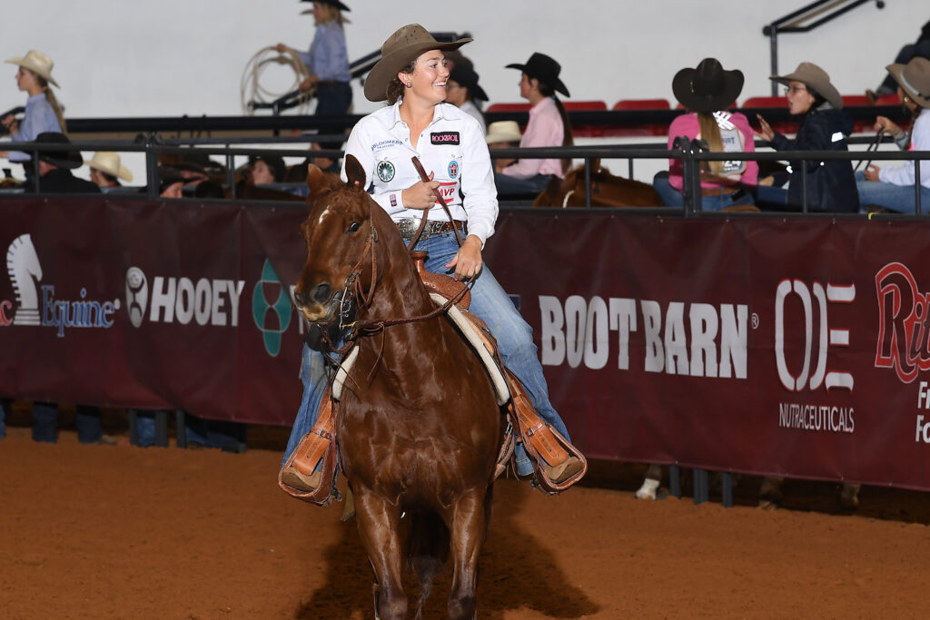 Rodeo competitor on brown horse riding past sponsor banners in arena during professional rodeo event