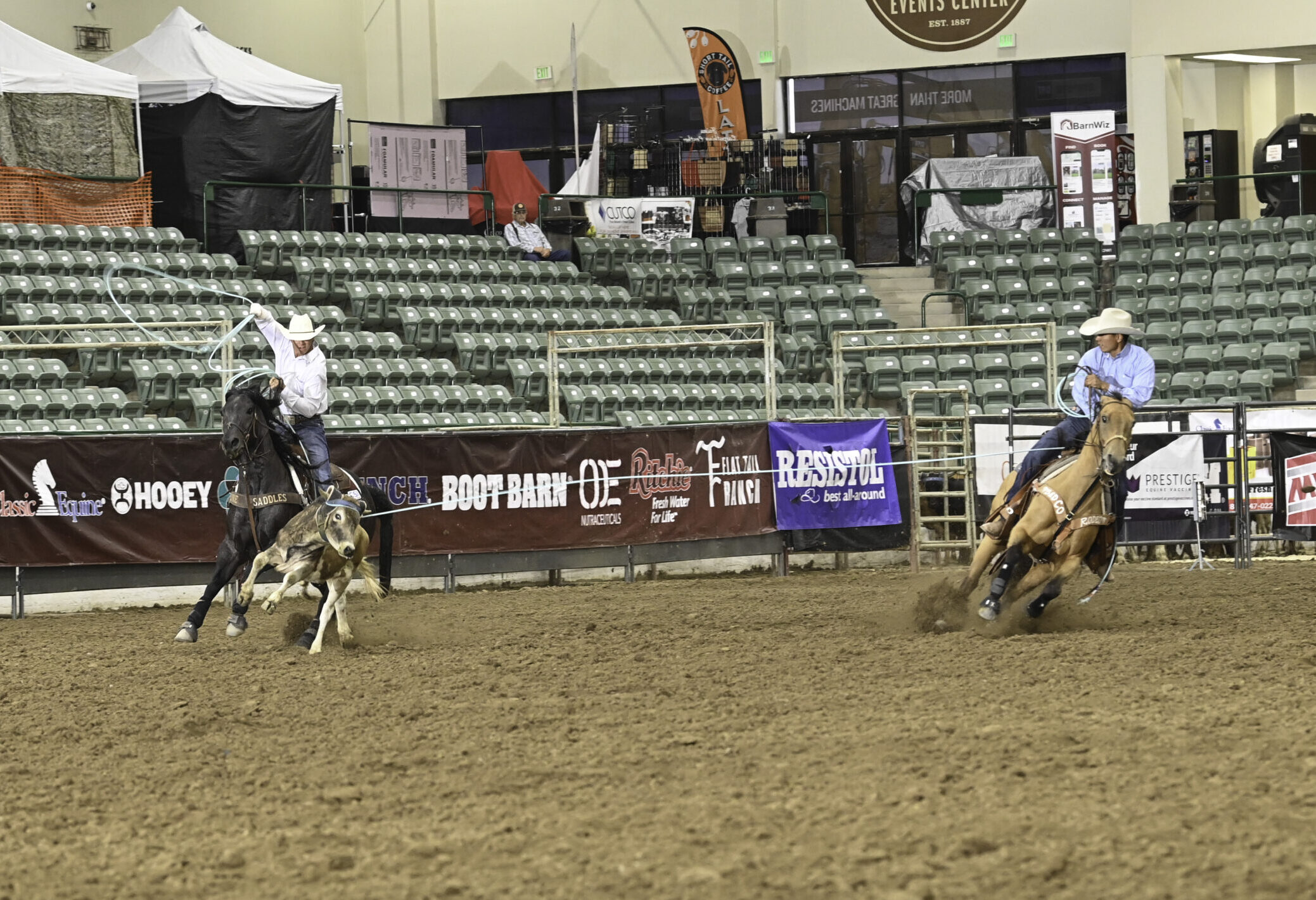 Two cowboys on horseback compete in a ranch sorting event indoors, with one rider steering a calf while the other waits nearby