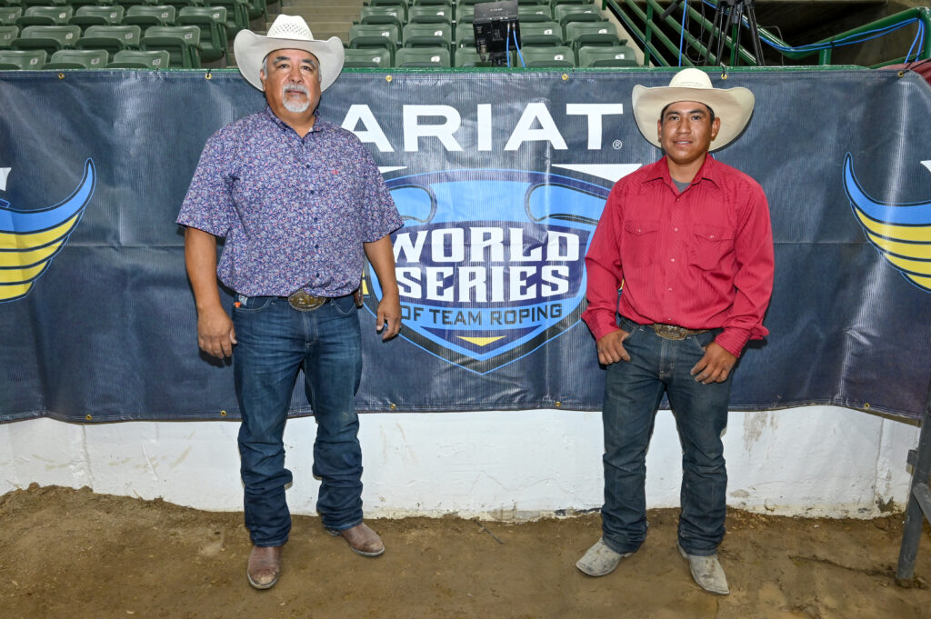Two men in cowboy hats and western shirts stand in front an Ariat World Series of Team Roping banner at an indoor arena