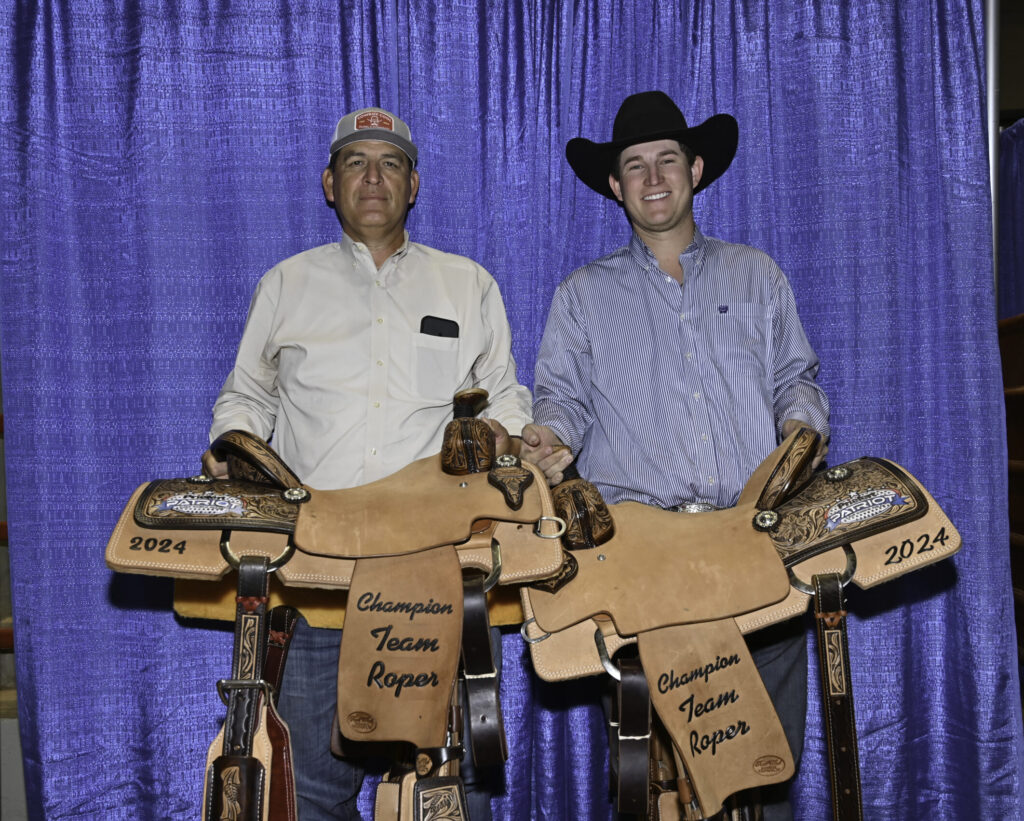 Two men with decorated saddles labeled "Champion Team Roper" and dated 2024 pose against a purple backdrop