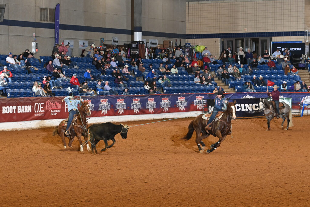 Cowboys on horseback work cattle during a ranch sorting competition at an indoor arena with spectators in the stands