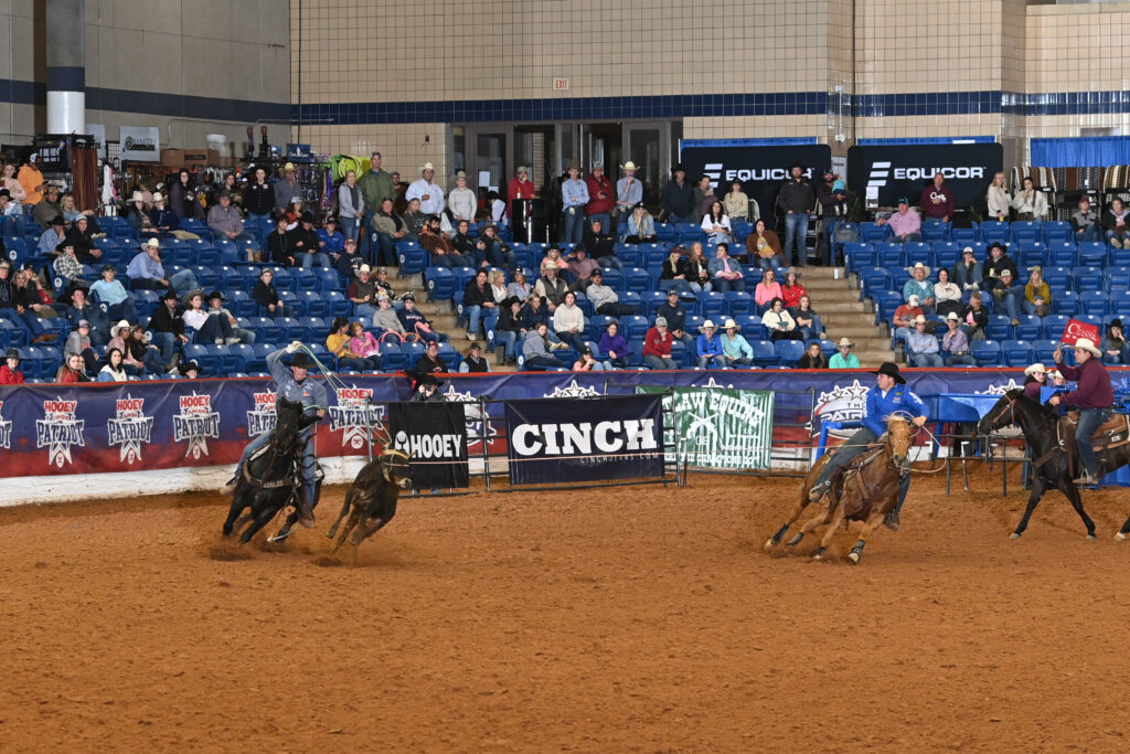 Cowboys on horseback separate cattle during a ranch sorting competition in an indoor arena with blue bleachers and sponsor signage