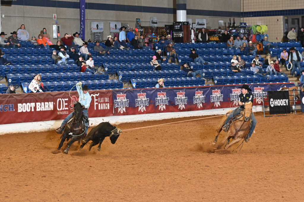 Two cowboys on horses compete in steer roping at an indoor rodeo arena with spectators in blue seats lining the venue