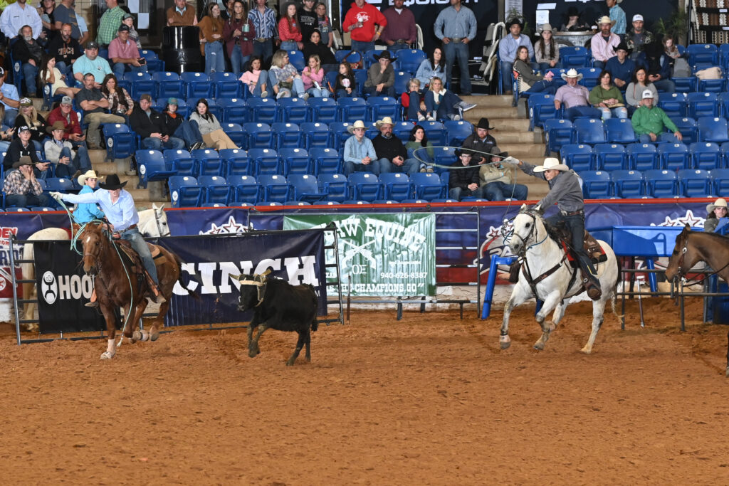 Cowboys on horseback herd a black steer across a dirt arena at a professional rodeo event with spectators in blue seats