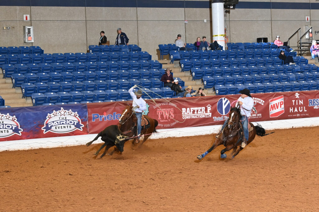 Two cowboys on horseback compete in a roping event at an indoor rodeo arena with blue seats and sponsor signage