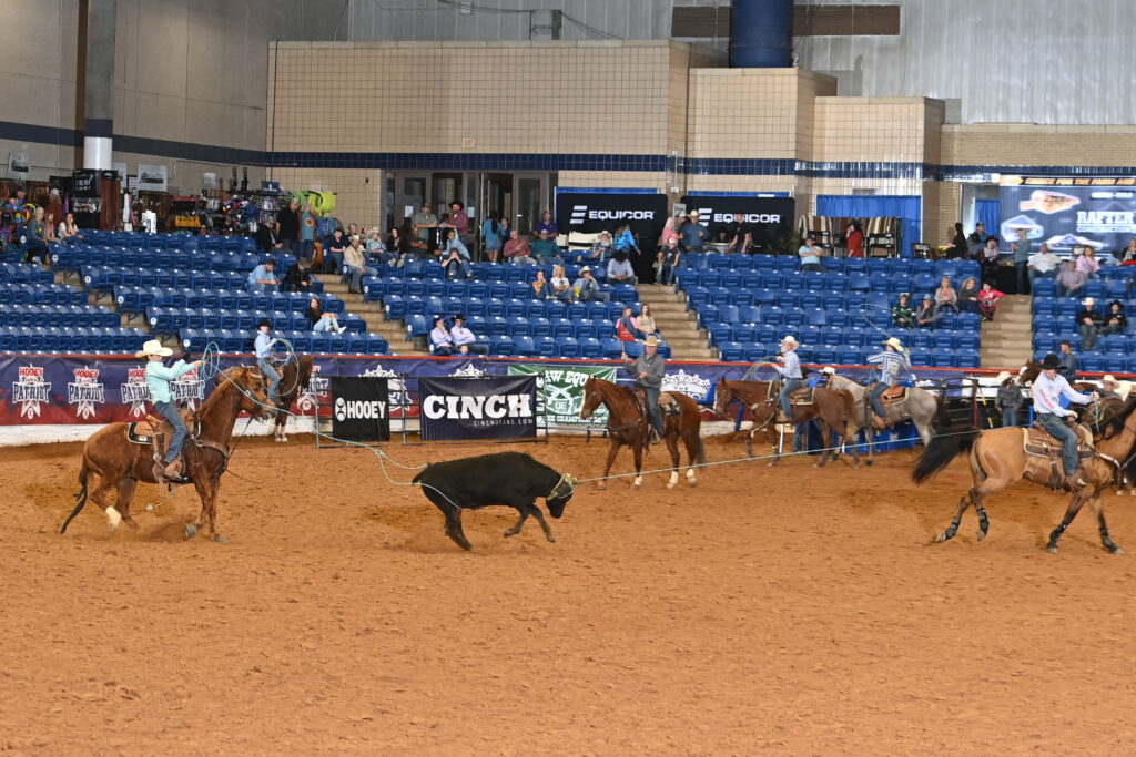 Steer roping competitors on horseback chase a black steer across the dirt arena at an indoor rodeo venue with blue bleachers and Cinch sponsorship signage