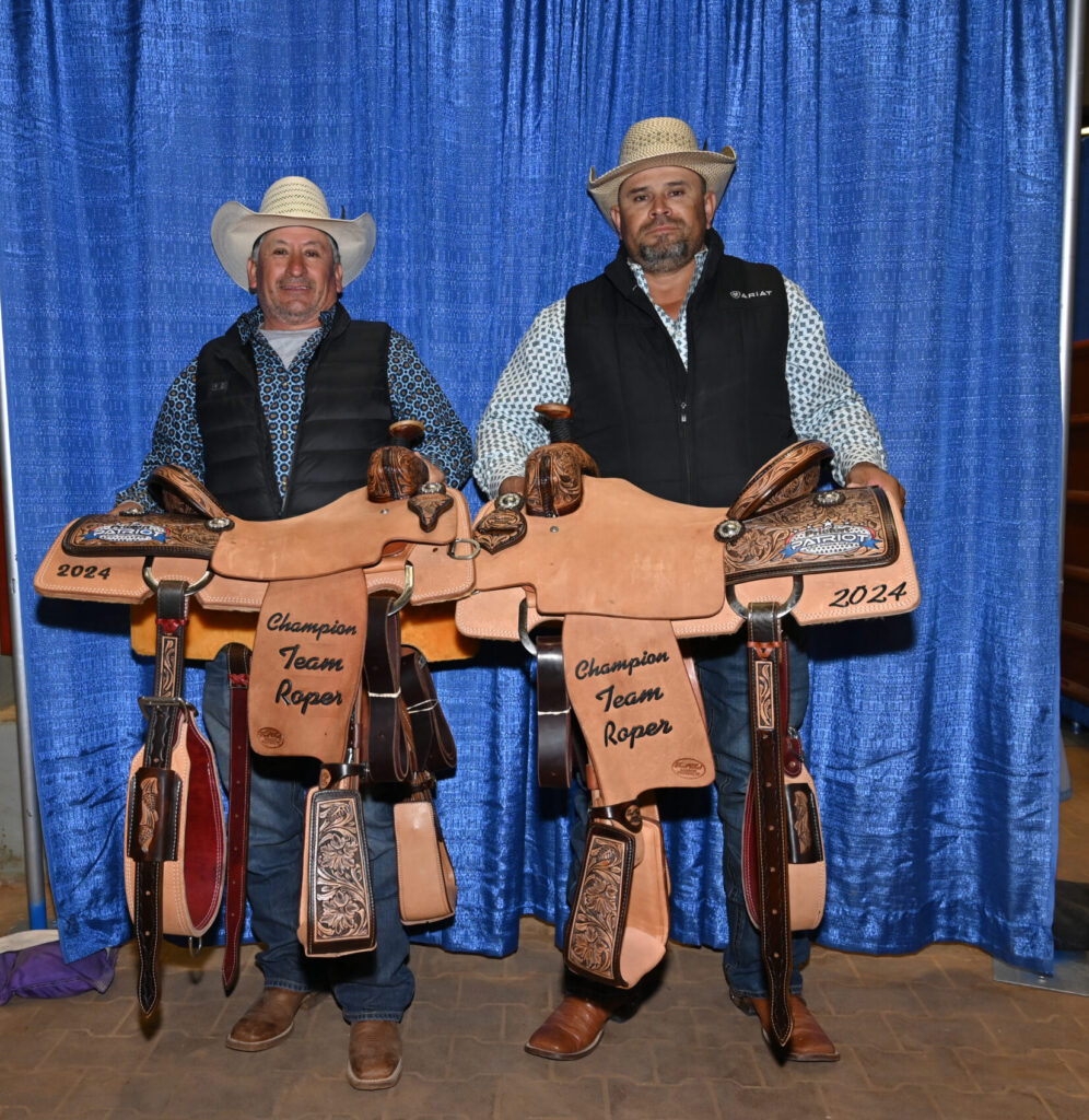 Two men in cowboy hats and western attire display ornate leather saddles labeled "Champion Team Roper 2024" against a blue backdrop