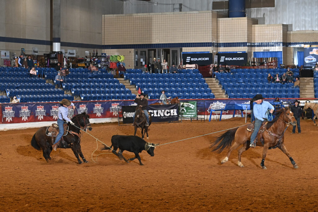 Cowboys on horseback rope a steer during a ranch sorting competition in an indoor arena with blue stadium seating