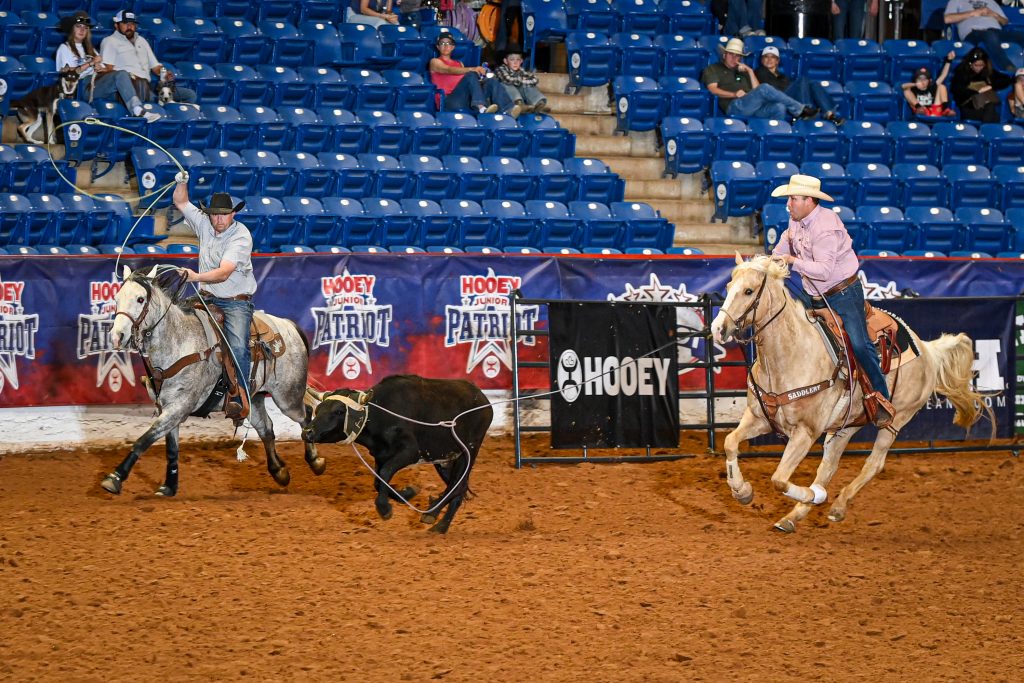 Two cowboys on horseback work together to rope a black steer during a rodeo event at an indoor arena with blue seats and sponsor signage