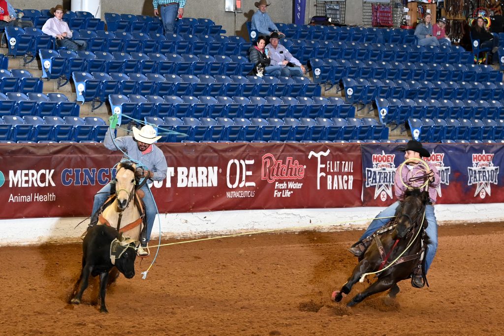 Cowboy roping steer during ranch sorting competition in indoor arena with blue stadium seating and sponsor signage visible
