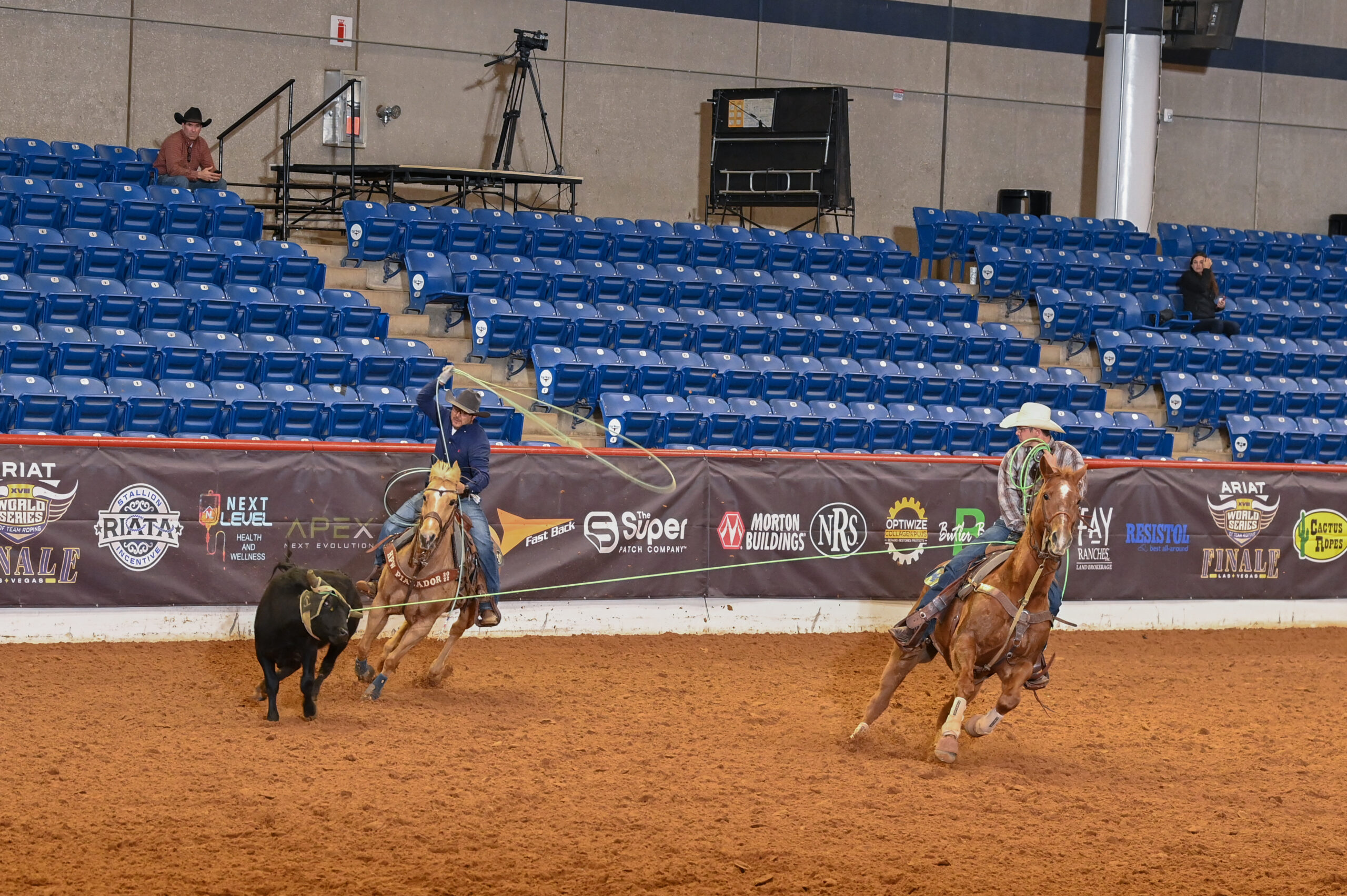 Two cowboys on horseback work to separate a black calf during a ranch sorting competition in an indoor arena with blue stadium seating