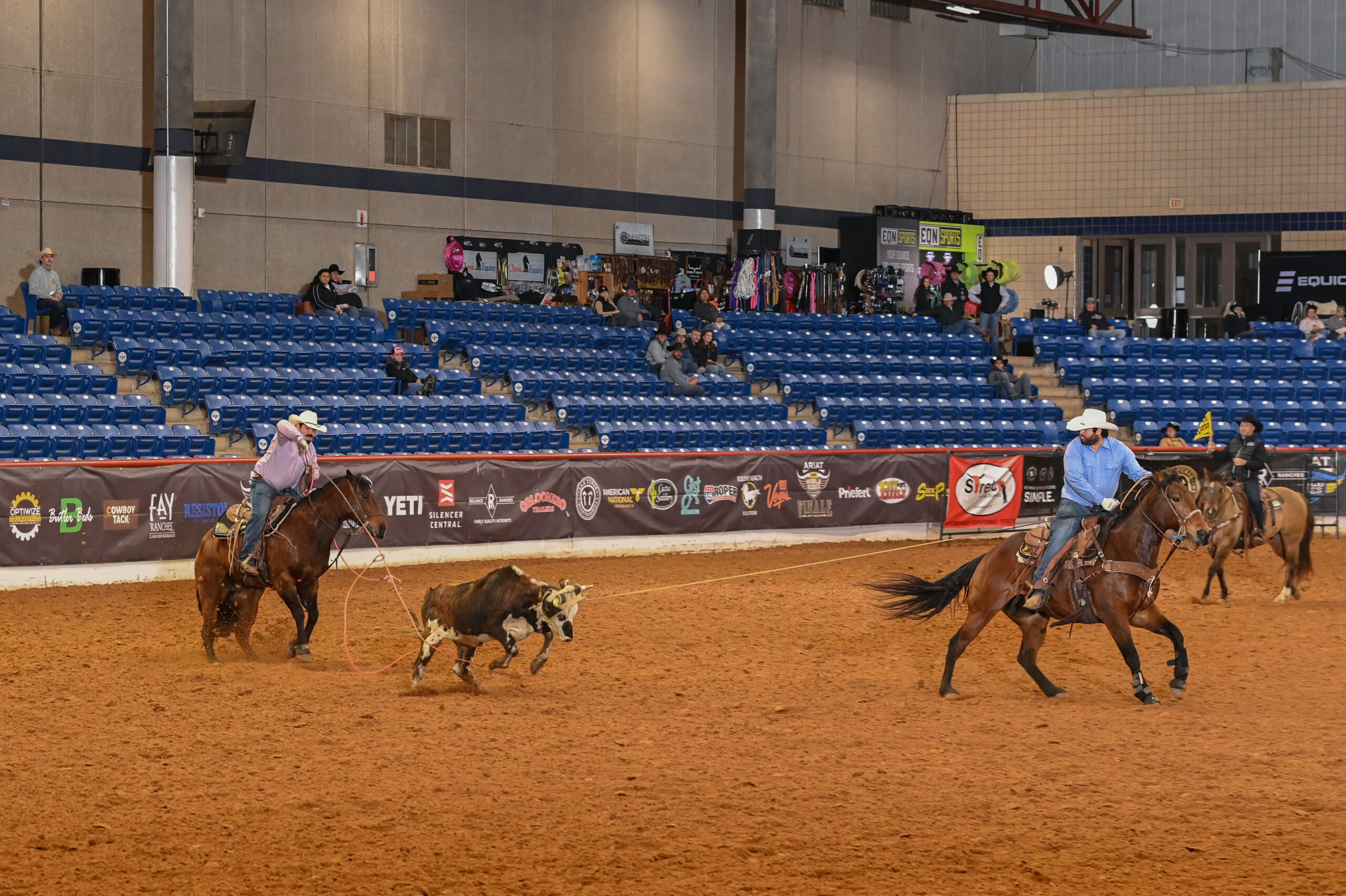 Two cowboys on horseback rope a steer during a ranch sorting competition in an indoor arena with blue bleacher seating
