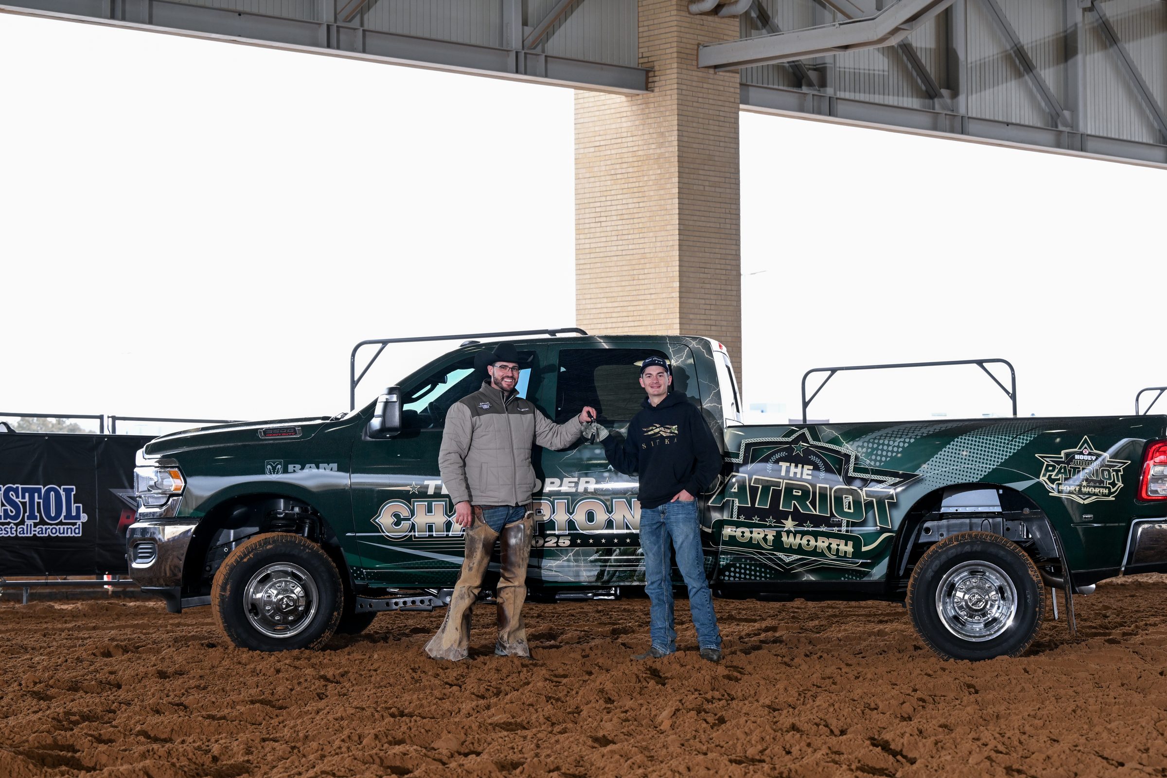 Two men stand between two green pickup trucks with rodeo branding at what appears to be a Fort Worth arena venue