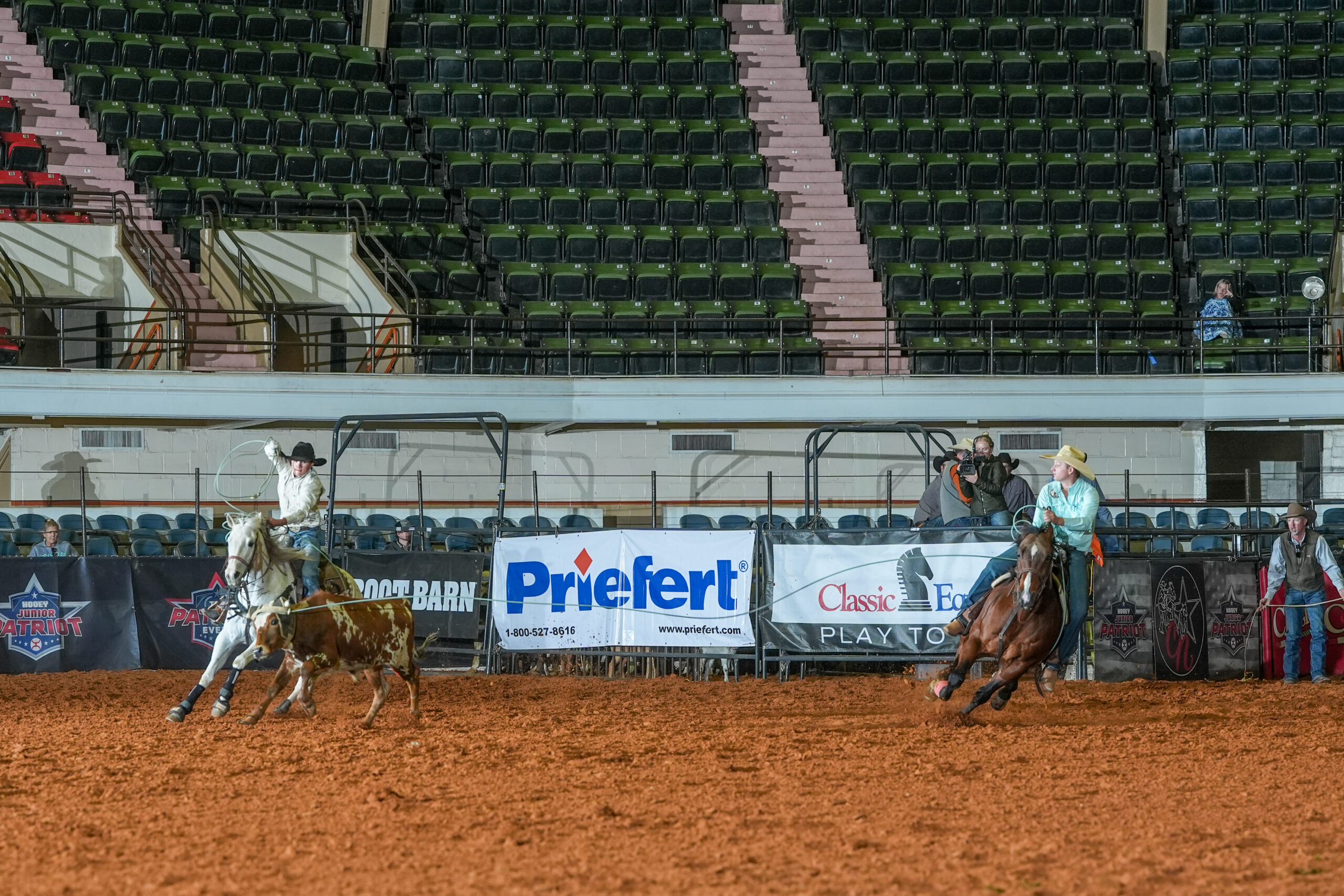 Rodeo competitor on horseback chases steer across arena dirt at indoor stadium with sparse crowd seating