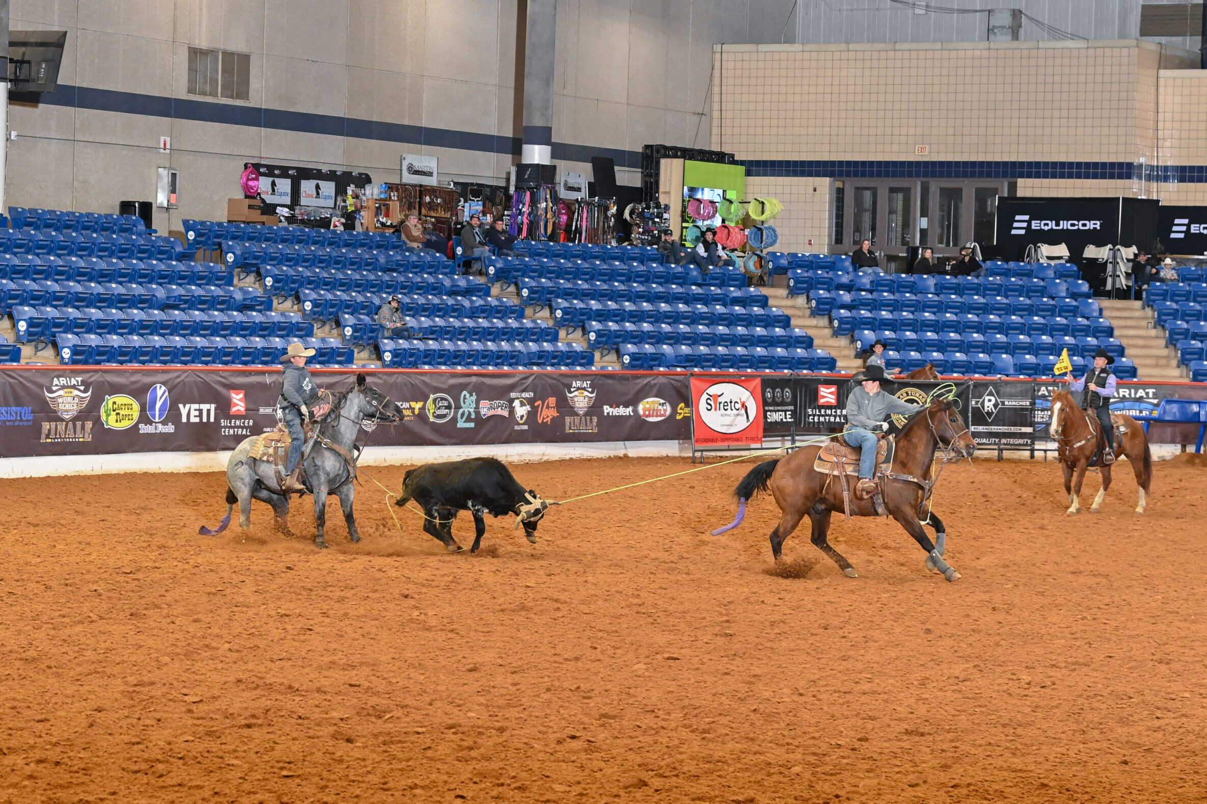 Cowboys on horseback work to separate a black steer during a ranch sorting competition in an indoor arena with blue stadium seating