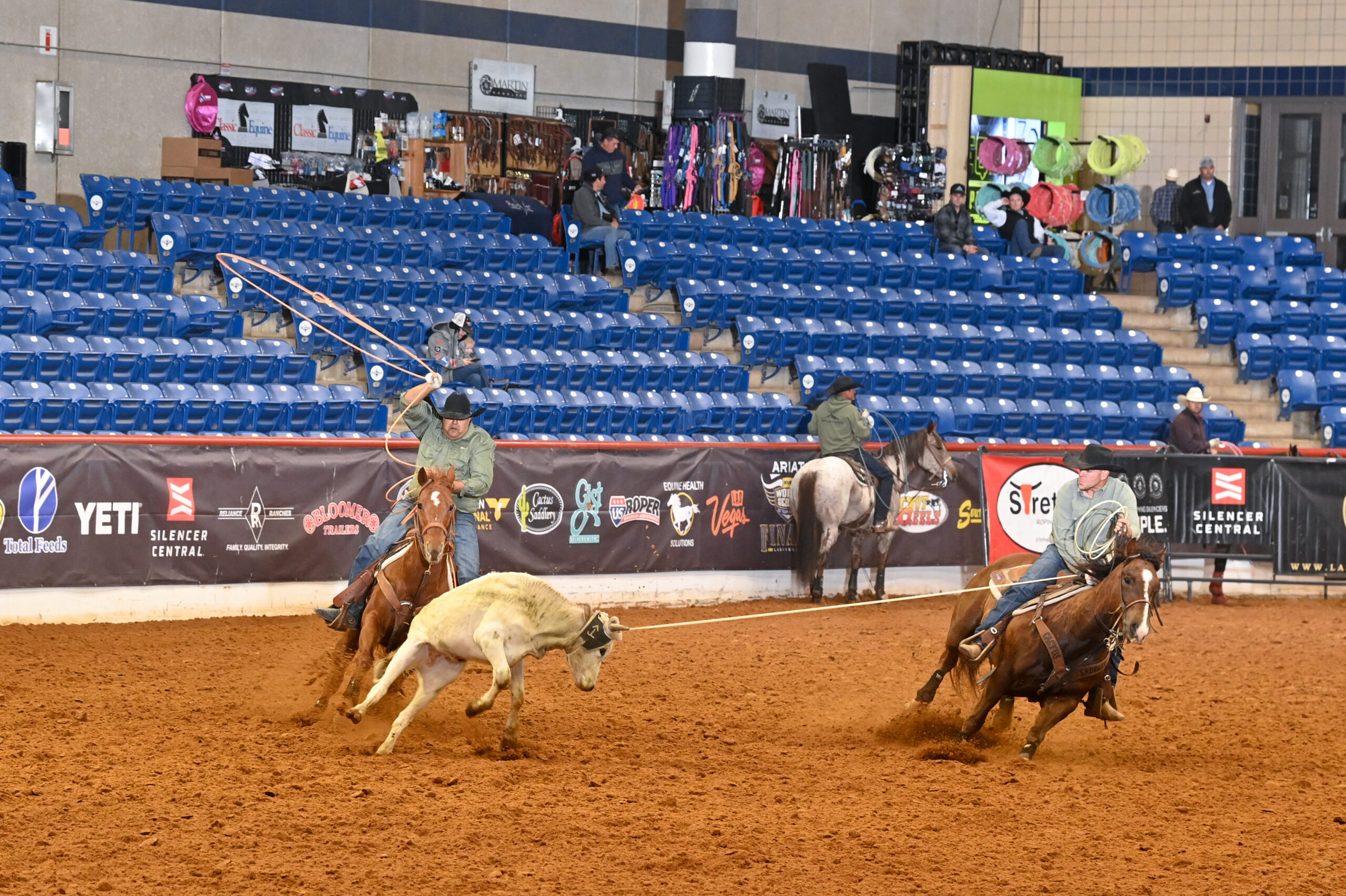 Rodeo competitor on horseback ropes a steer during an indoor arena event, with spectators seated in blue chairs behind the advertising barrier