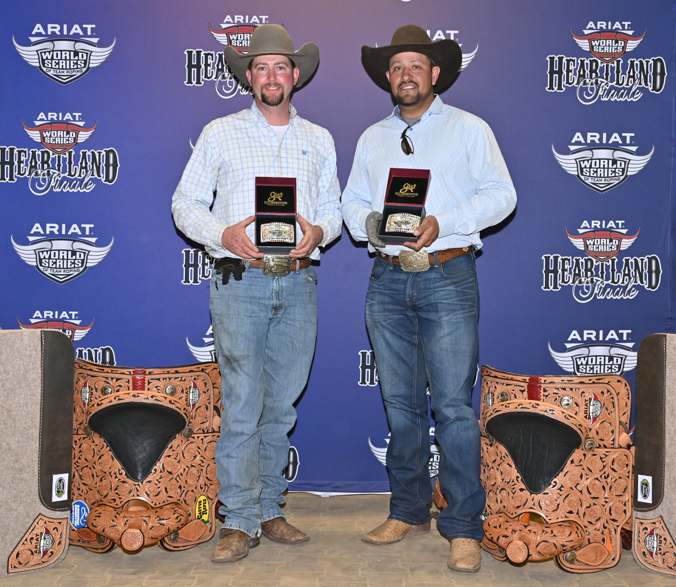 Two cowboys hold championship buckles and trophies at the ARIAT World Series Heartland Finale event