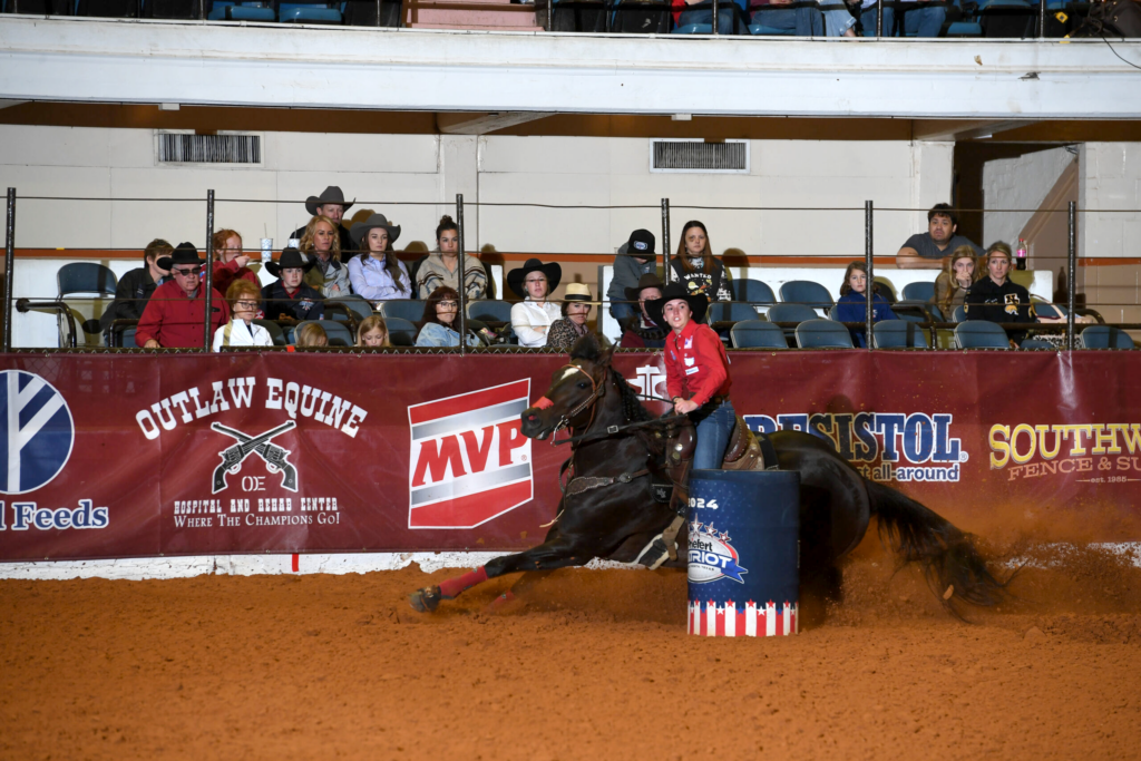 Barrel racer on dark horse rounds a barrel during rodeo competition as spectators watch from stands behind advertising banners